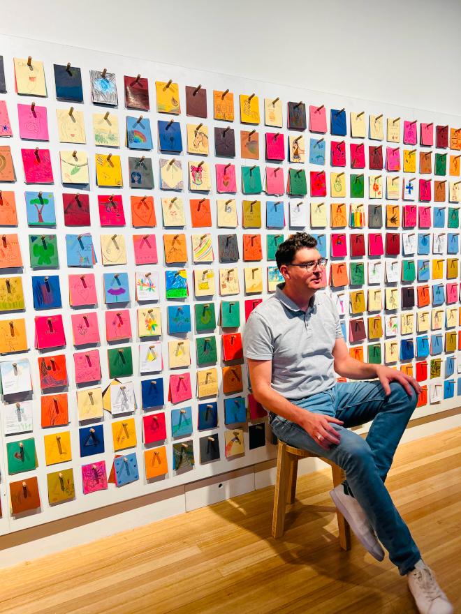 Dallas with colorful paper behind him, sitting at an exhibition at Te Papa Museum in Wellington.